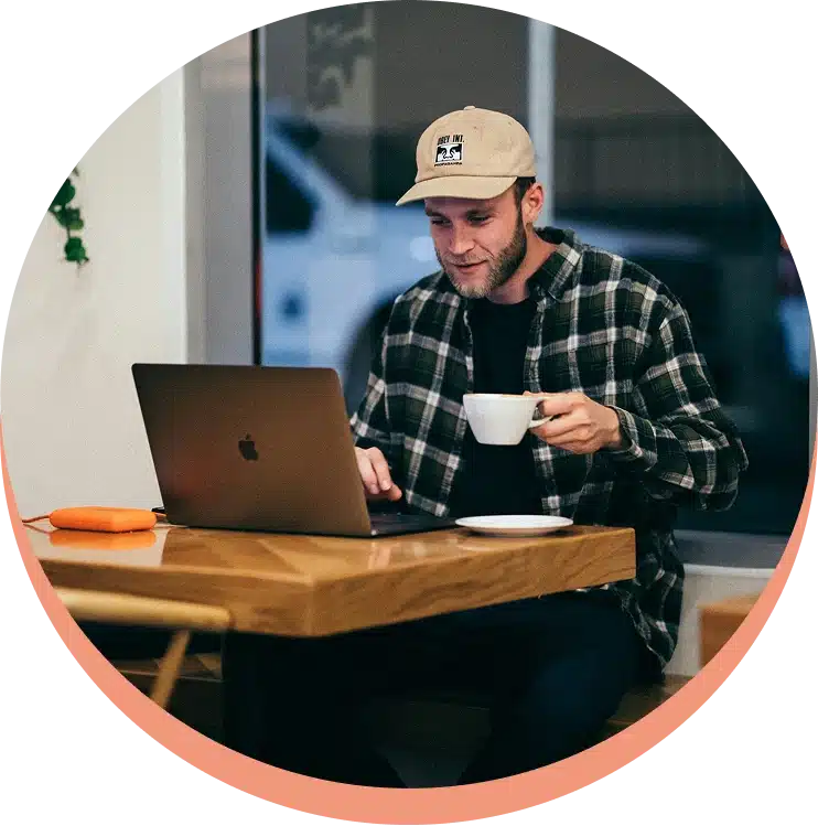 A man in a baseball cap works on his laptop at a cafe, holding a cup of coffee.