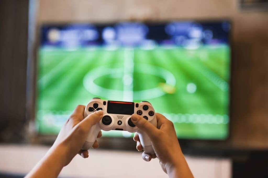 Hands holding a game controller in front of a blurred soccer video game screen, symbolizing technology addiction and anxiety.